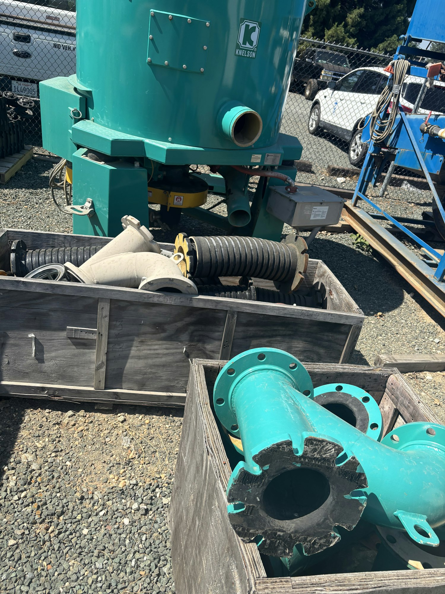 The image shows industrial equipment, including a large green machine and various pipes and fittings in wooden crates.