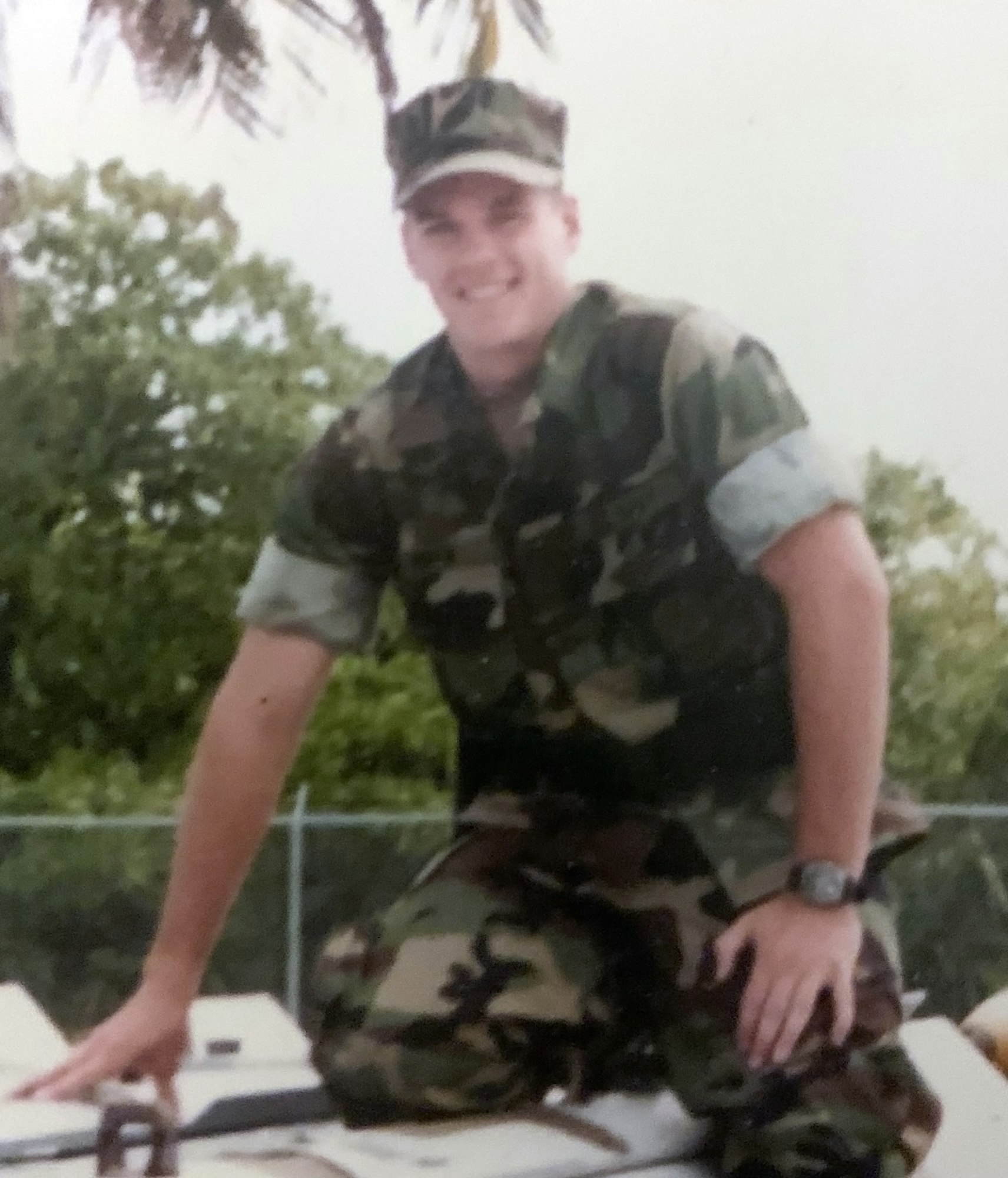 A person in military camouflage with a cap, smiling, outdoors with palm trees in the background.