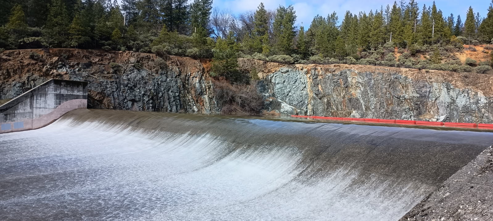 A dam with water flowing over it, surrounded by rocky cliffs and dense trees.