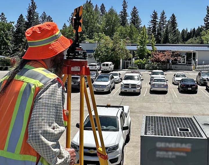 A person in a safety vest uses surveying equipment in a parking lot near a building and trees.