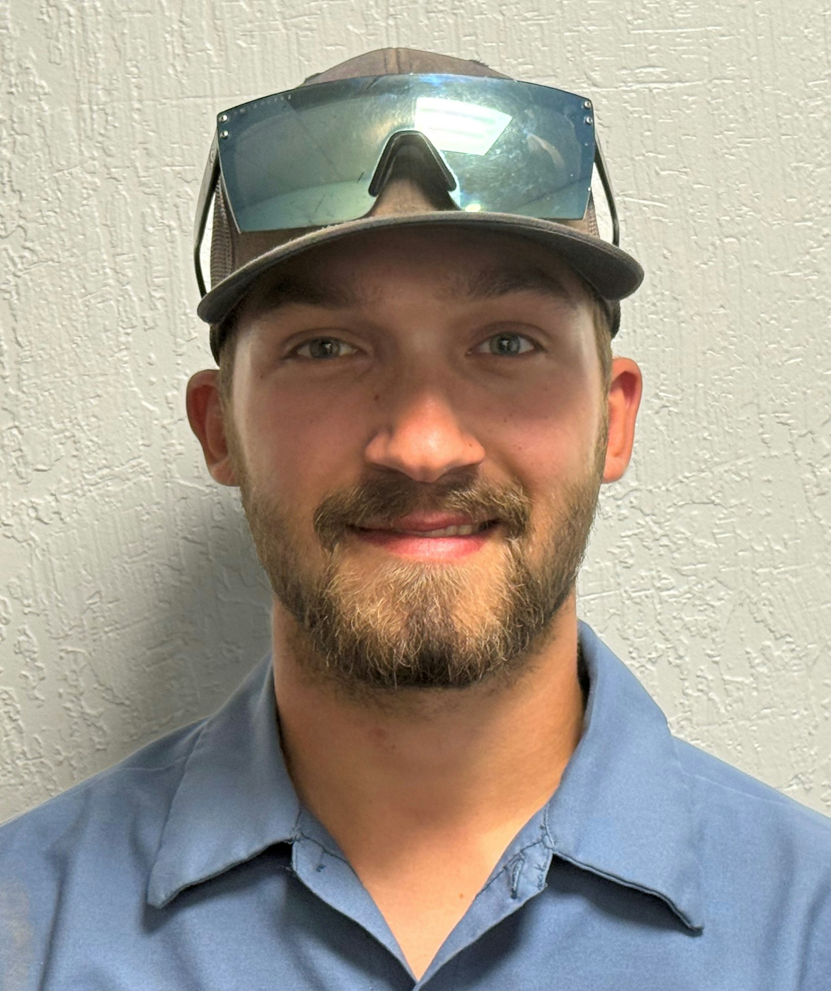 A man wearing a hat and sunglasses on his head, smiling, with a textured wall in the background.