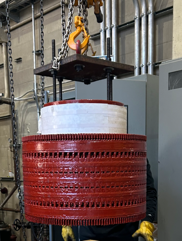 A large, red cylindrical industrial component being lifted by a hoist in a workshop.