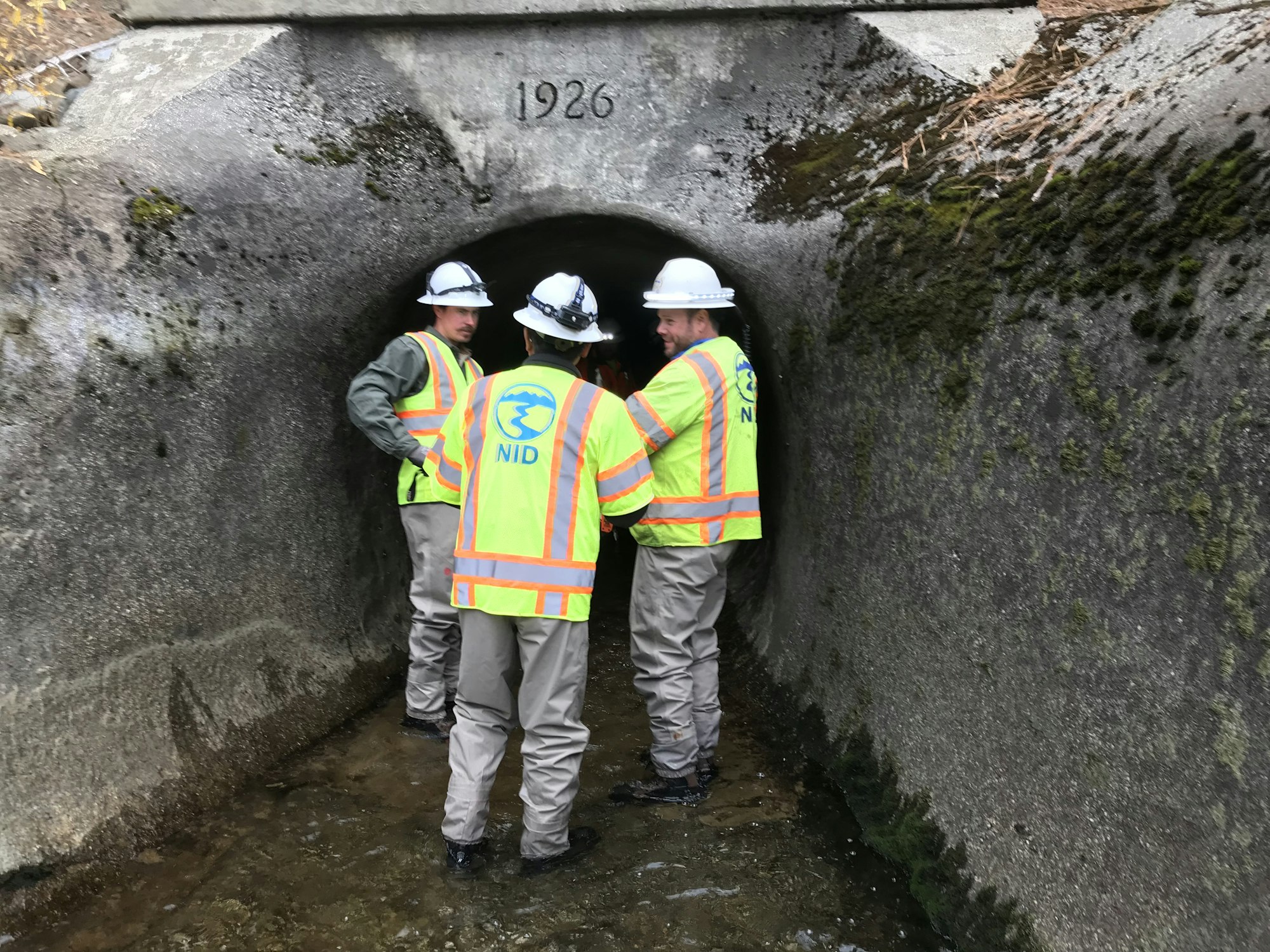 Three workers in safety gear are inspecting a tunnel marked "1926," likely related to water or infrastructure maintenance.