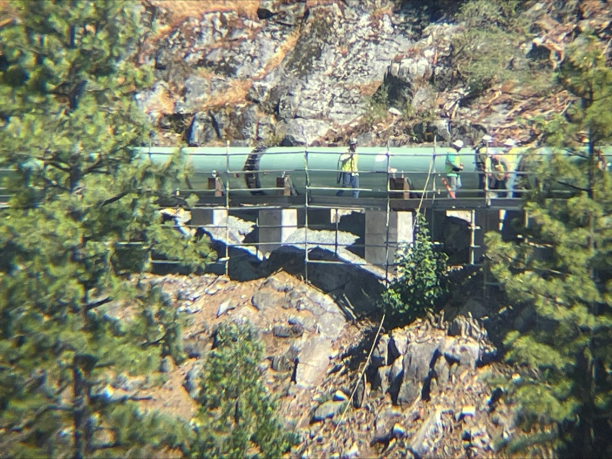 Large pipeline on a bridge surrounded by trees with people inspecting the site.