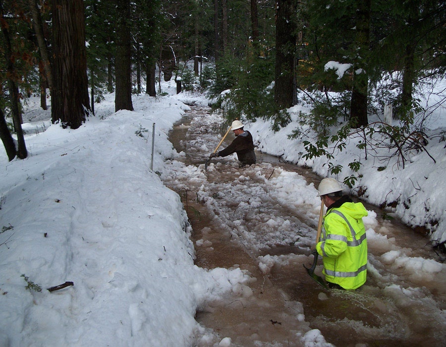 Two workers in snow-covered forest clear a flooded path, using shovels in muddy water.