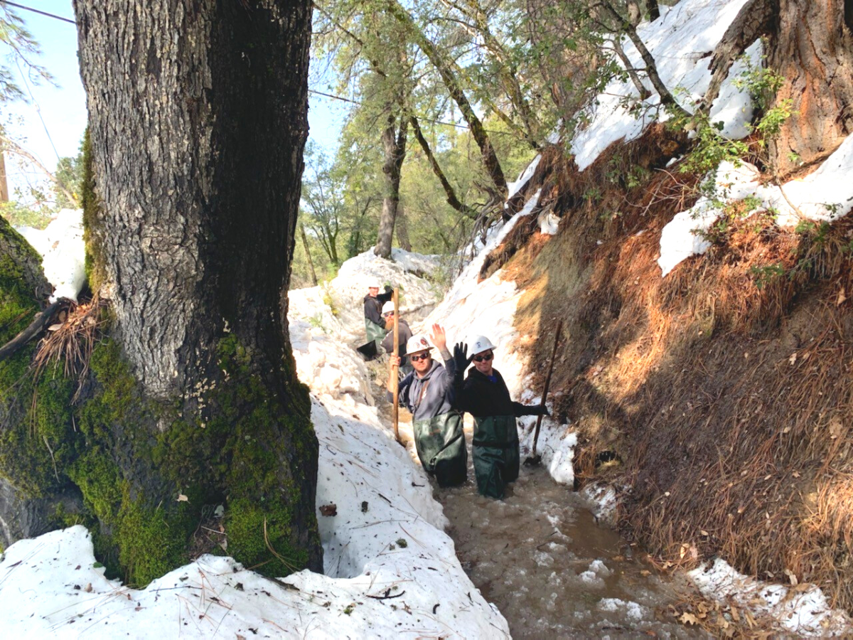 Two people in waders wave while standing in a snowy, slushy area near trees, enjoying an outdoor adventure.