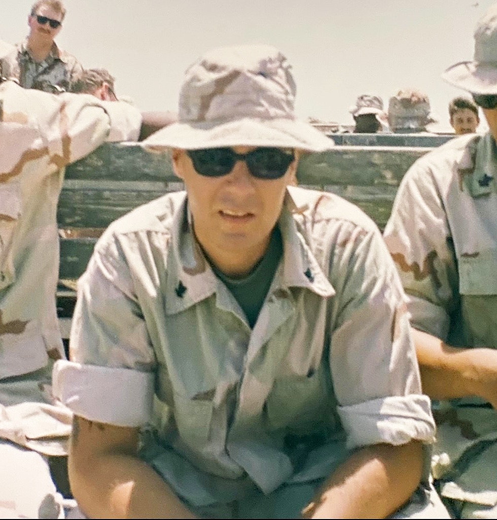 Person wearing camouflage and sunglasses with others in military attire in the background.