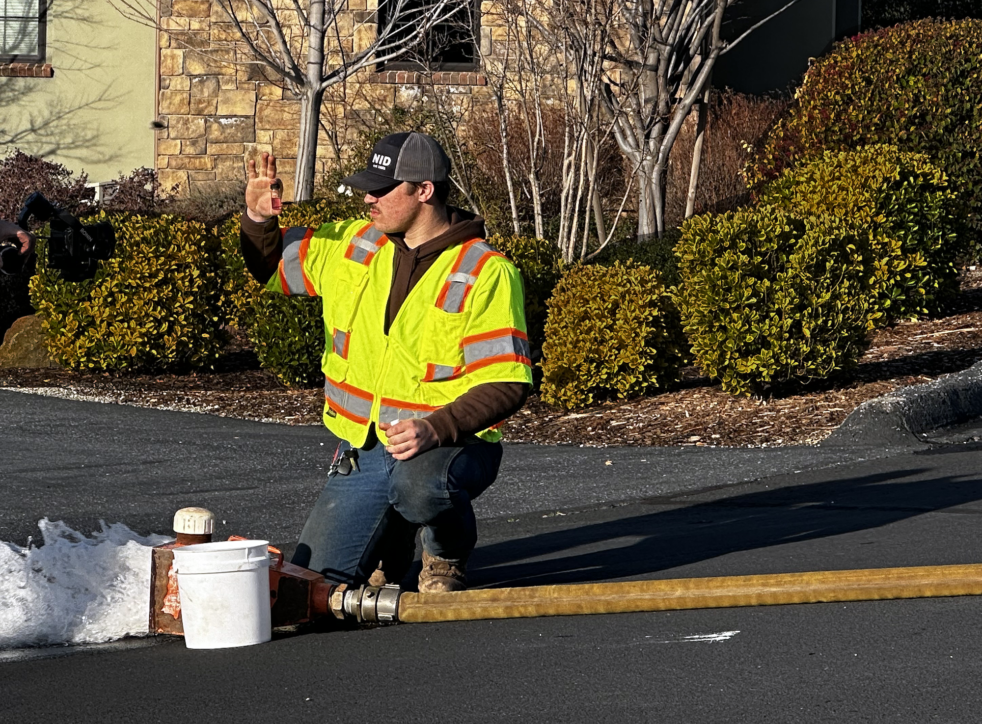 A worker in a high-visibility vest kneels by a hydrant, examining a water sample in a small jar.