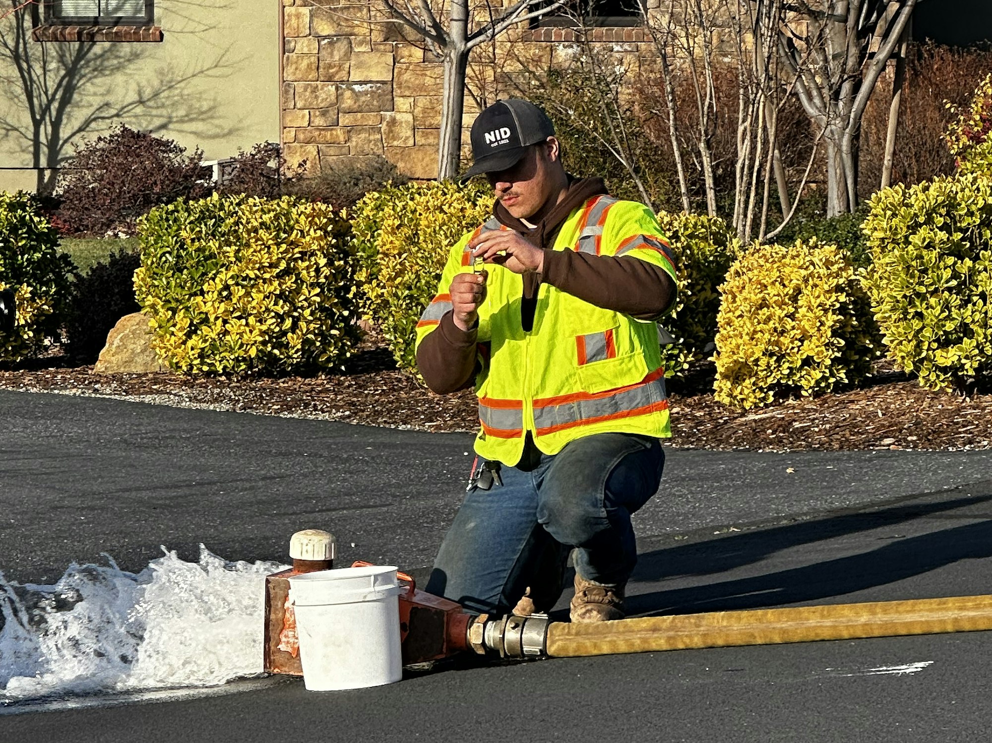 A worker in a safety vest kneels near a flowing fire hydrant, taking a sample or measurement with a small device.