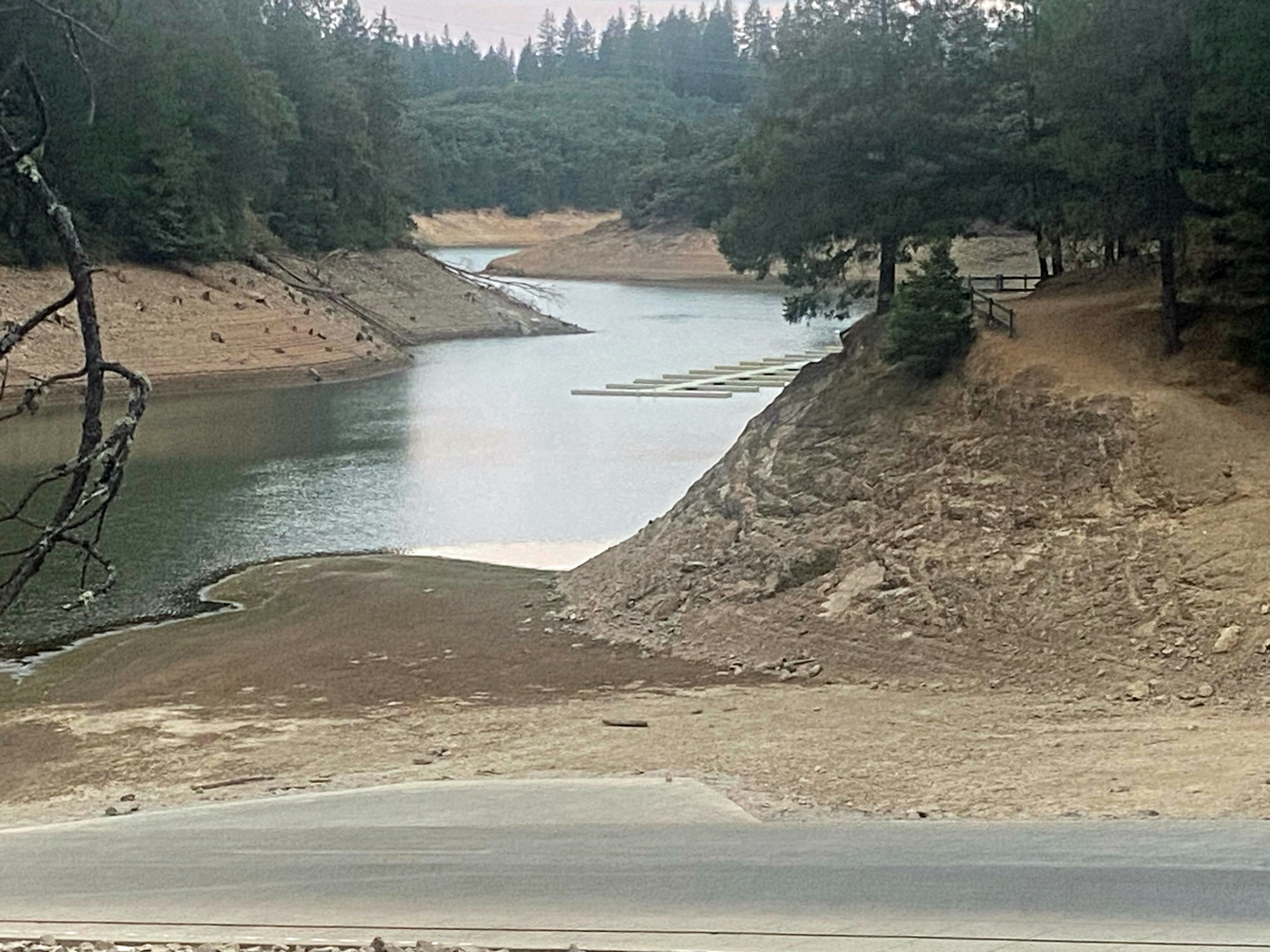 A depleted reservoir with exposed bank areas, trees, and a path along the ridge.