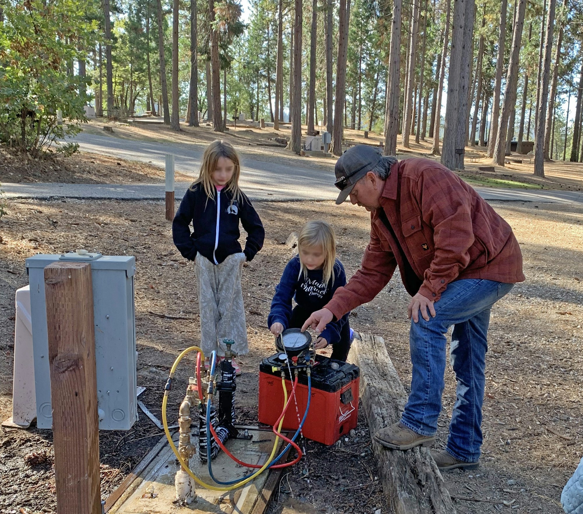 A man helps two girls with a setup involving gauges and hoses in a forested camping area.