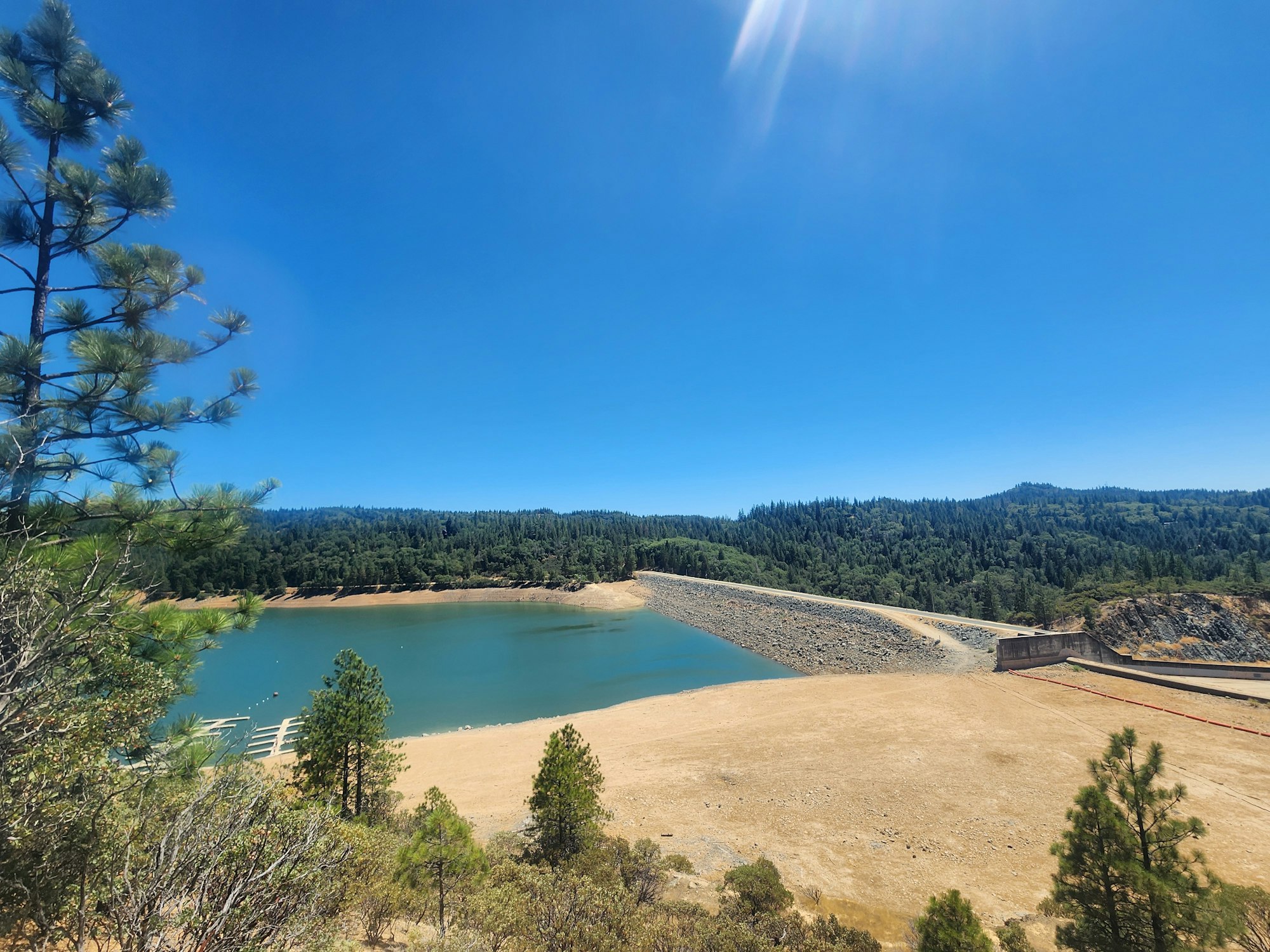 A clear day showing a reservoir, dam, pine trees, and surrounding forested hills under a blue sky.