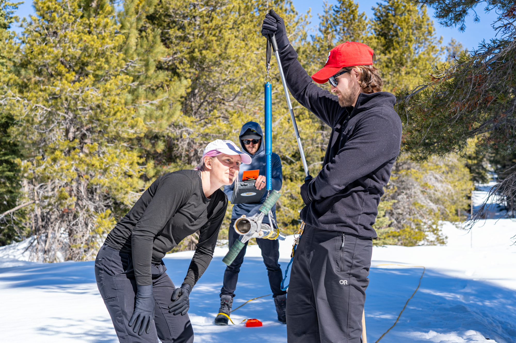 Three people measuring snow depth in a snowy forest with equipment.