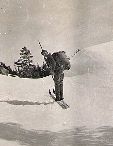 A person skiing uphill in snowy terrain, carrying a backpack and a rifle, with trees in the background.