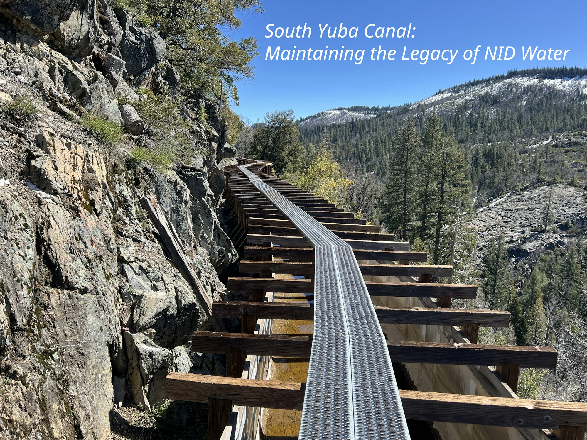 A flume on a wooden structure along a rocky hillside with trees and snow-capped mountains in the background, and text overlay about water legacy.