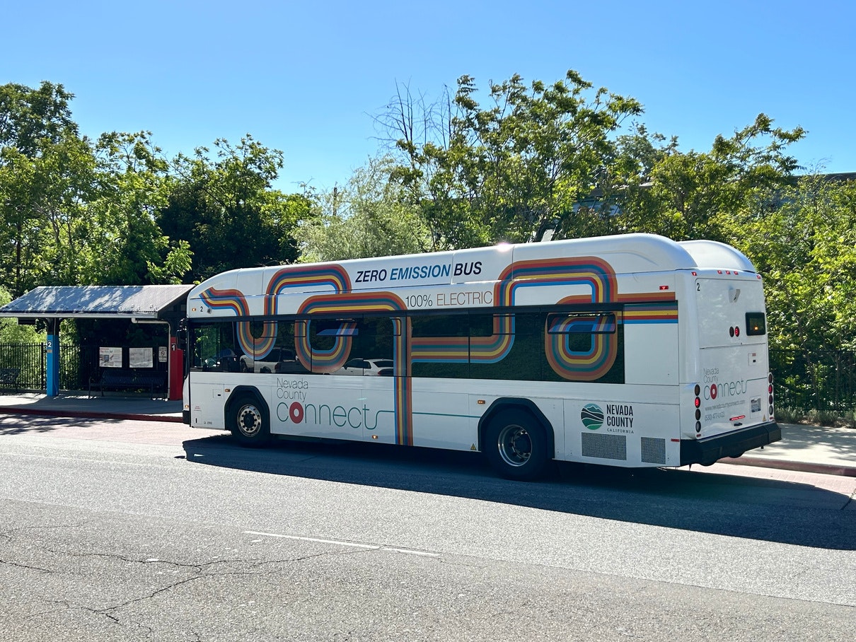 Electric zero-emission bus at a stop, labeled "Nevada County Connects," with trees and a shelter nearby.