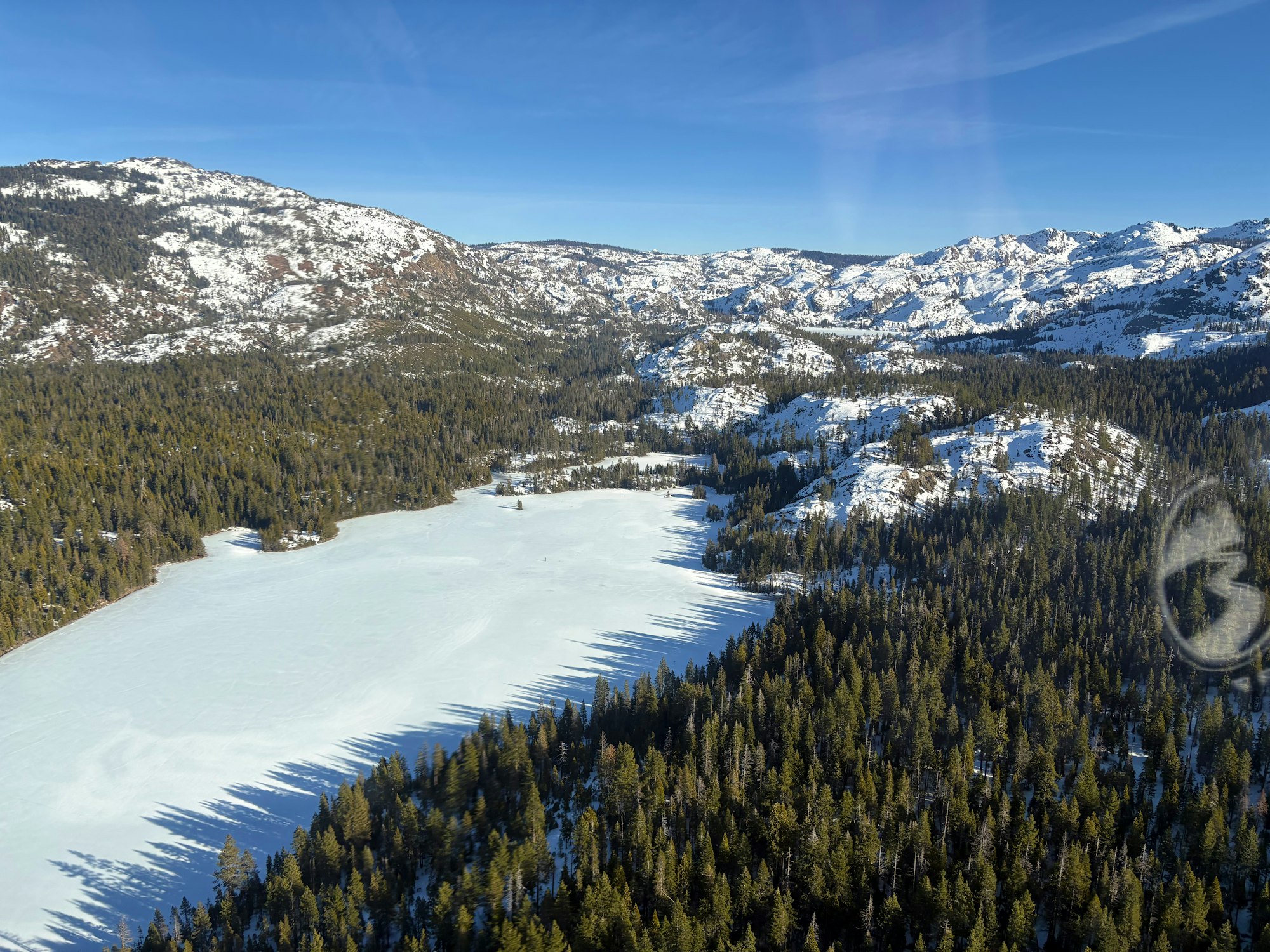 Aerial view of a snowy mountain landscape with a frozen lake surrounded by dense forests and a clear blue sky.