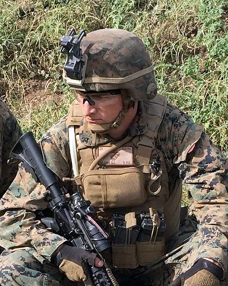 A soldier in camouflage with a helmet, goggles, and rifle, kneeling on grass.
