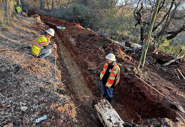 Construction workers in orange vests and hard hats standing by a freshly dug trench in a forested area.