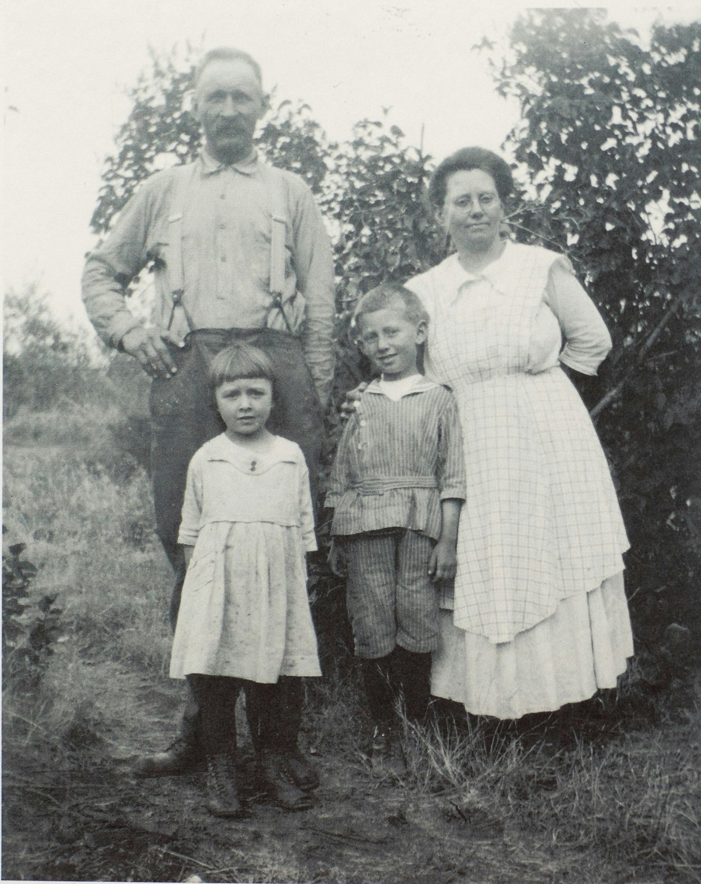 A black-and-white photo of a family: two adults and two children, standing outdoors among plants. The setting appears rural.