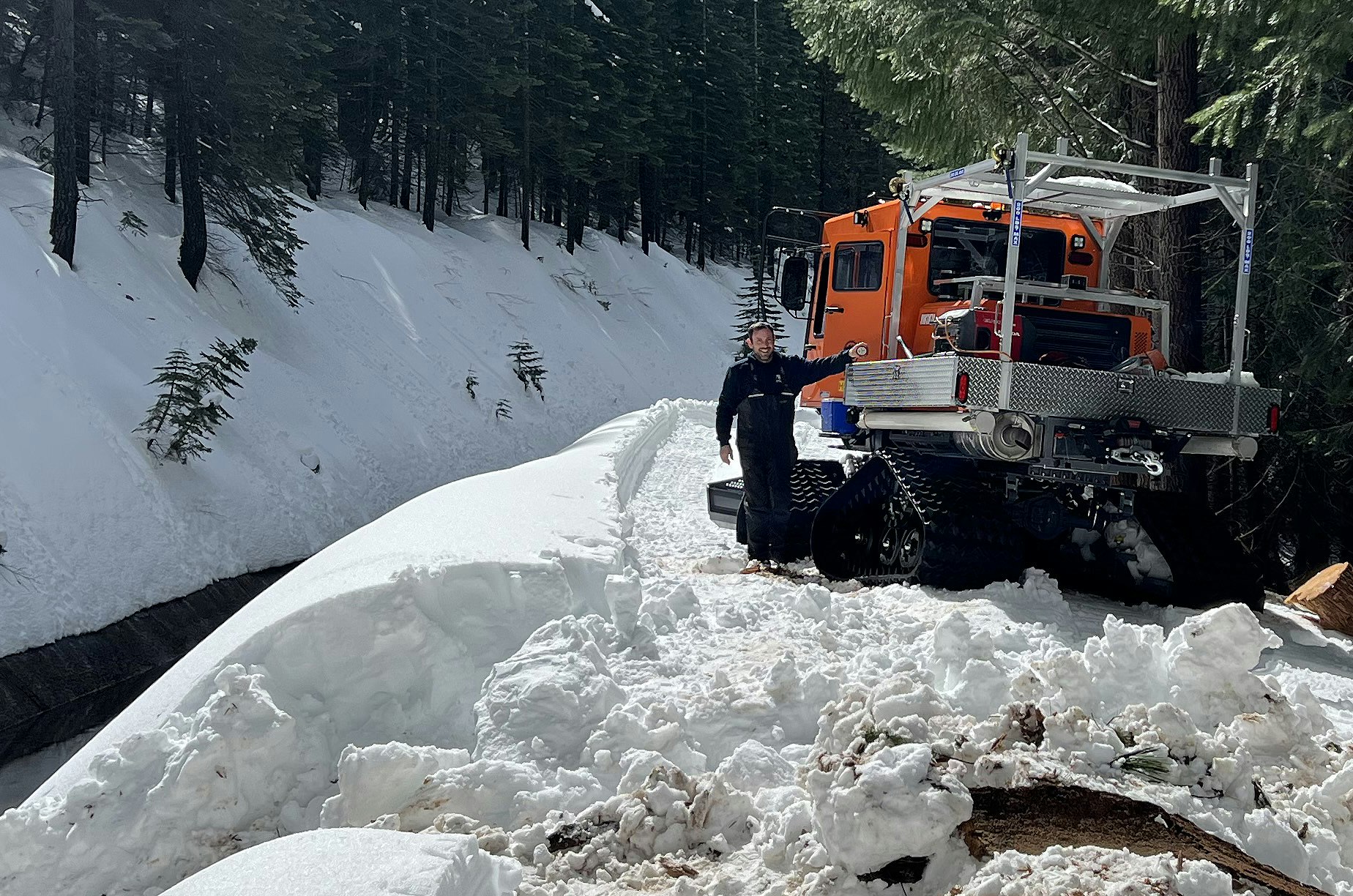 An orange snowcat vehicle and a person on a snowy forest road with cleared snow around.