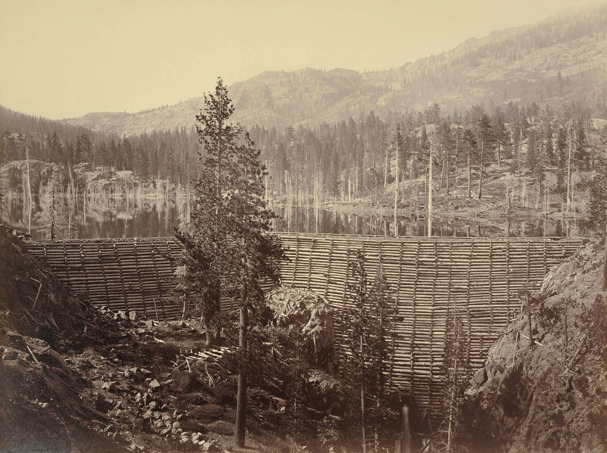 A log dam in a forested mountainous landscape with a lake in the background.