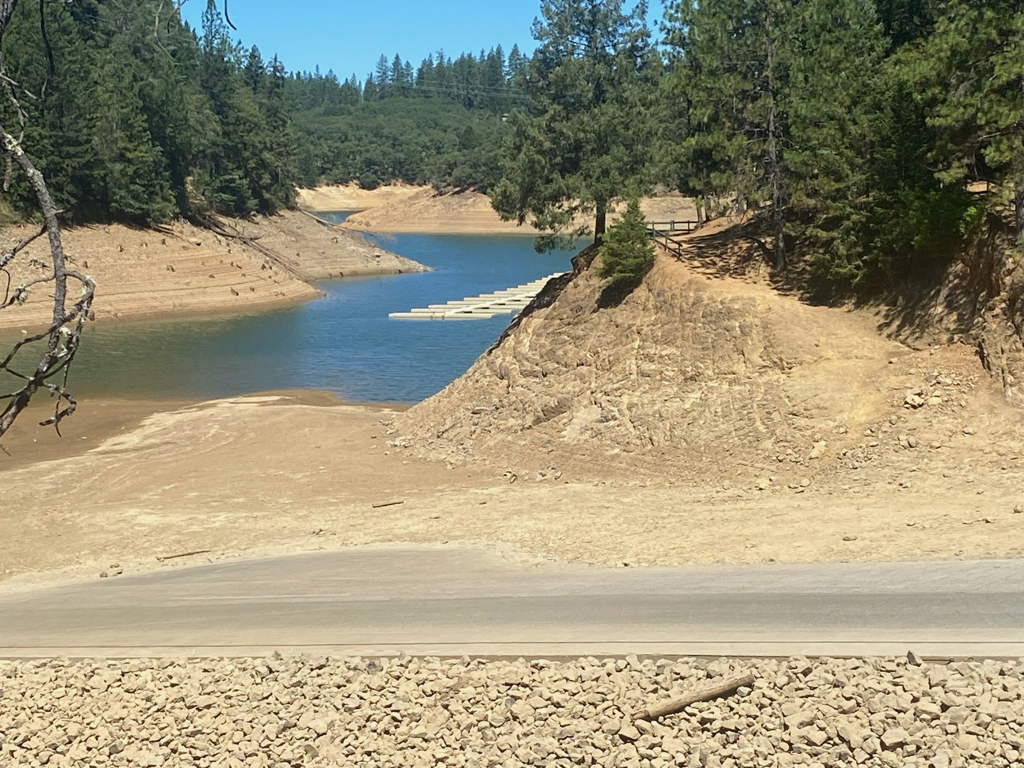 A reservoir with low water levels surrounded by trees and exposed banks, with a floating dock in the distance.