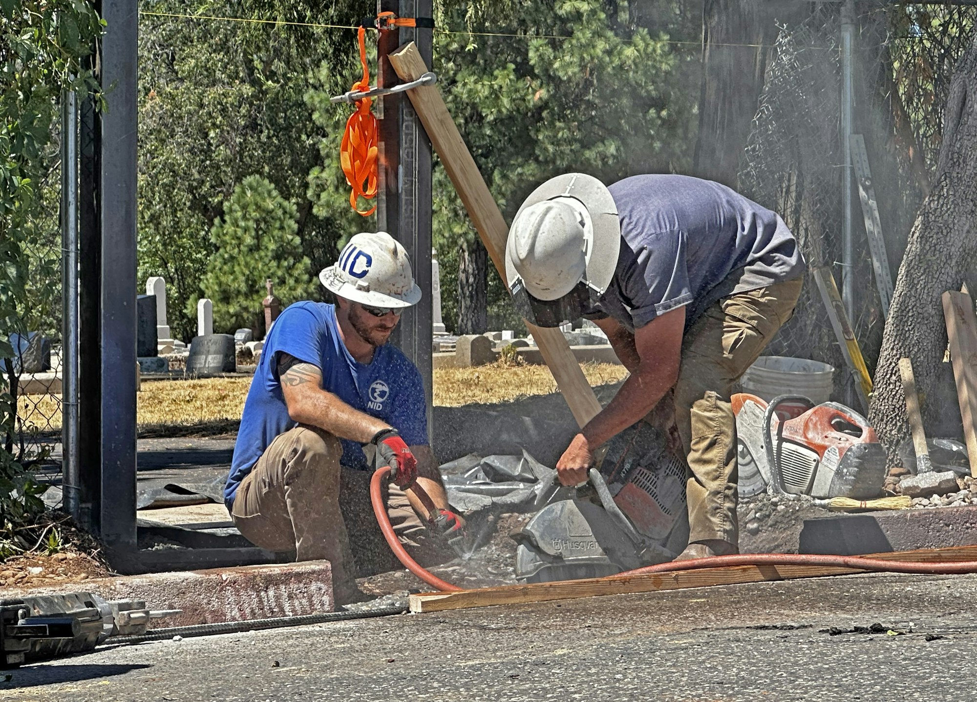 Two workers in hard hats are using construction tools outdoors. Dust and equipment are visible.