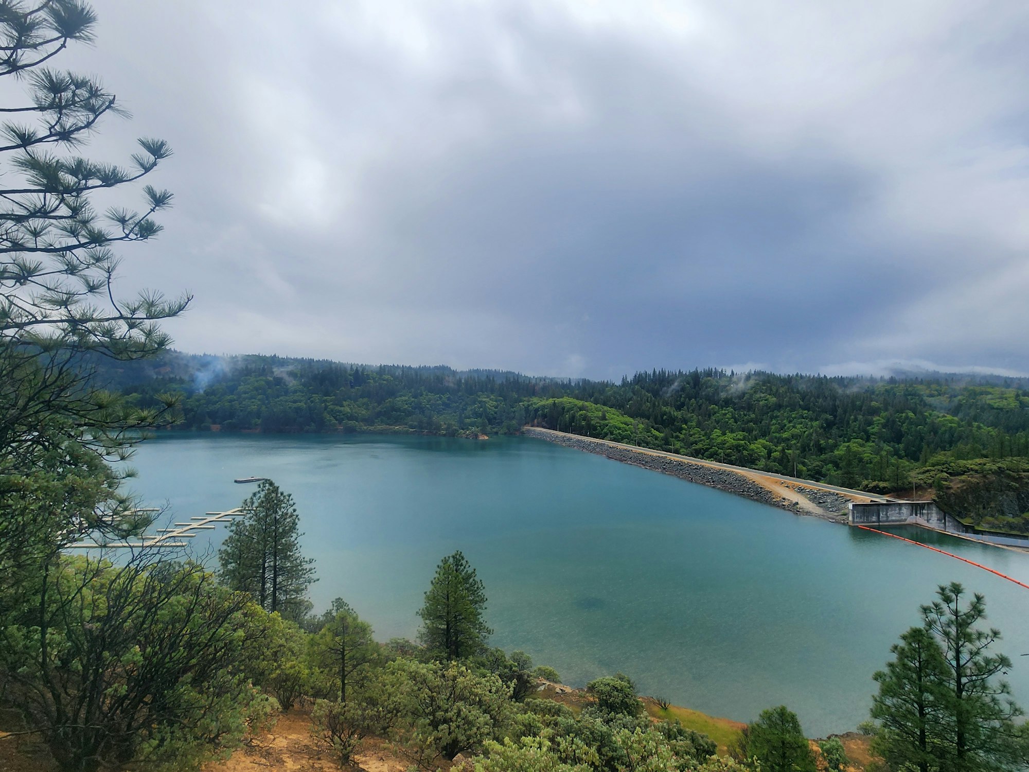 A serene lake with a dam, surrounded by forest under a cloudy sky.