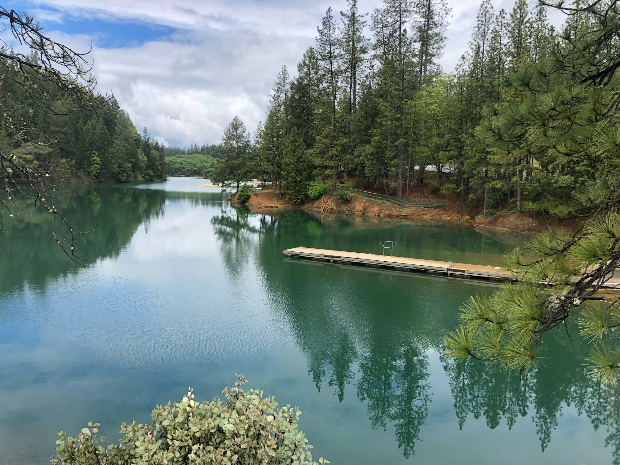 Serene lake with reflective water, dock, pine trees, and cloudy skies.
