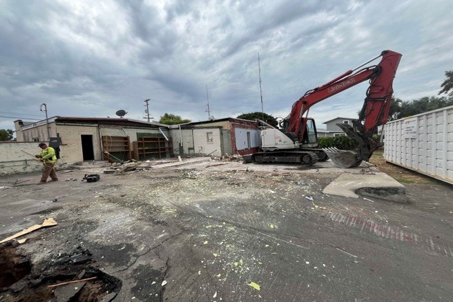 Demolition site with an excavator and a person wearing a high-visibility vest amidst debris and a partly demolished building.