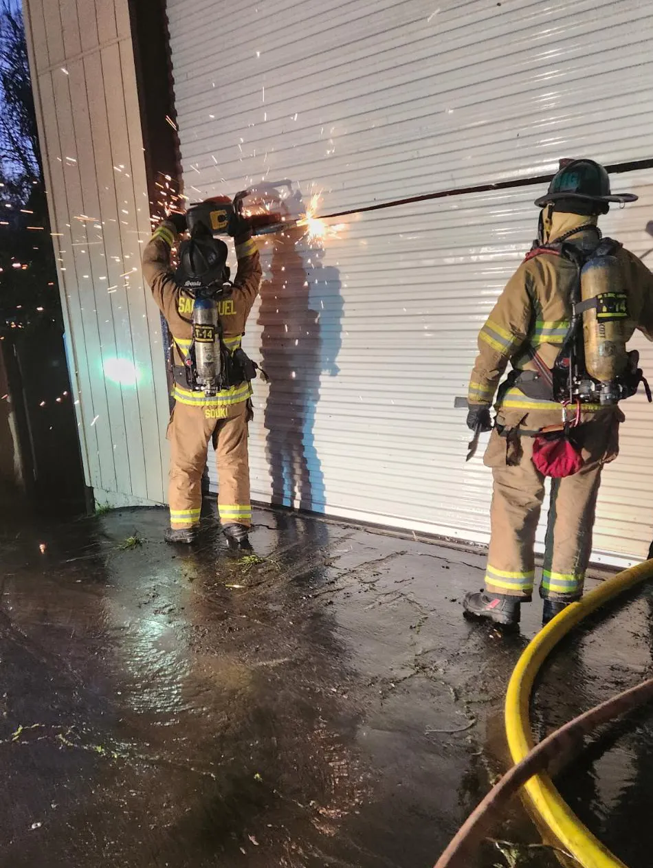 Two firefighters are cutting through a door with sparks flying, while another firefighter observes what’s happening.