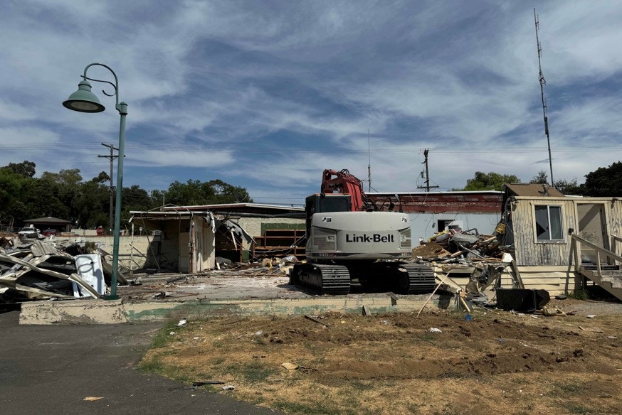A Link-Belt excavator near a demolished building with debris scattered around under a clear sky.