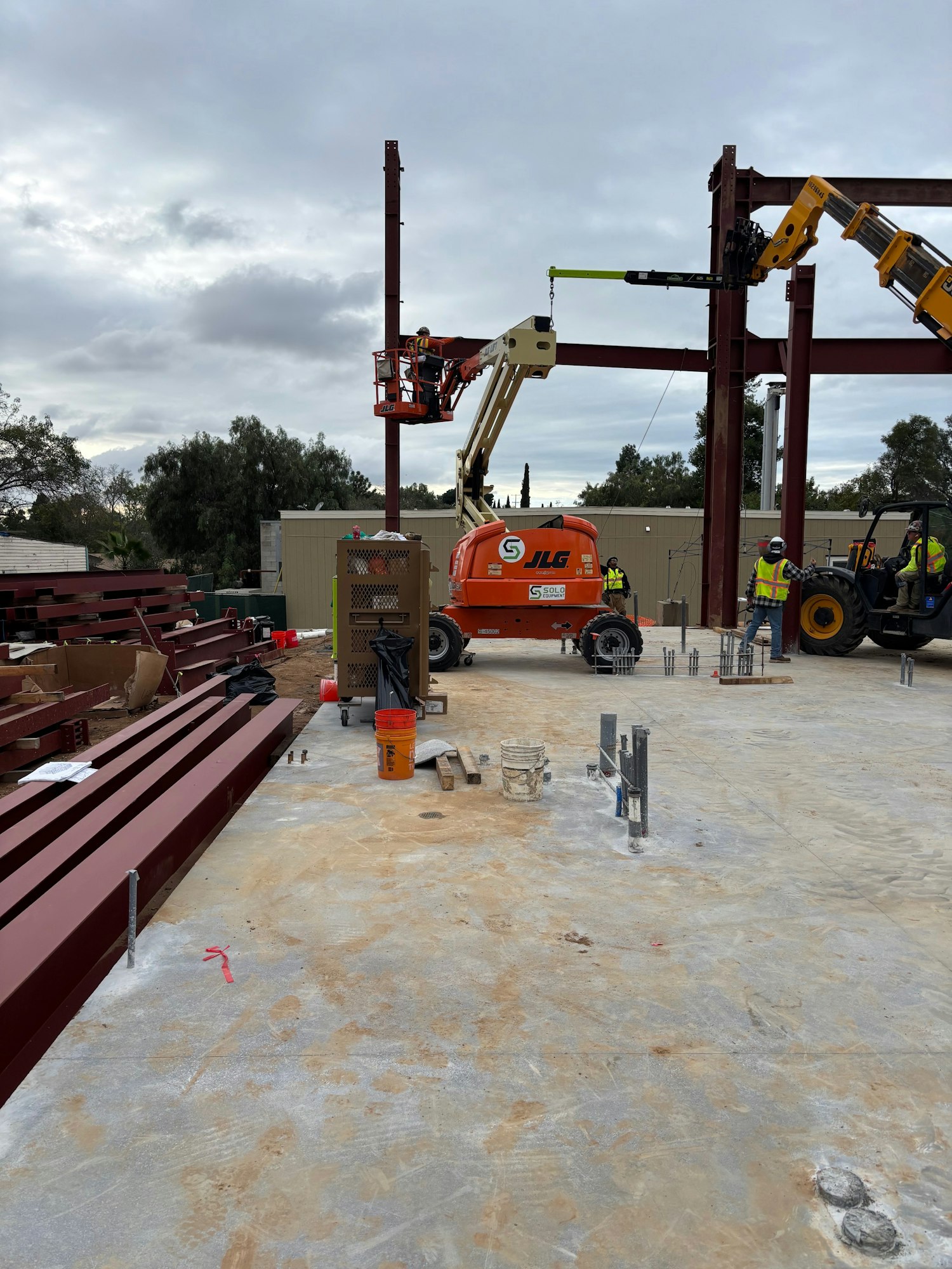 The image shows a construction site with workers, cranes, and steel beams. Equipment is set up for building work.