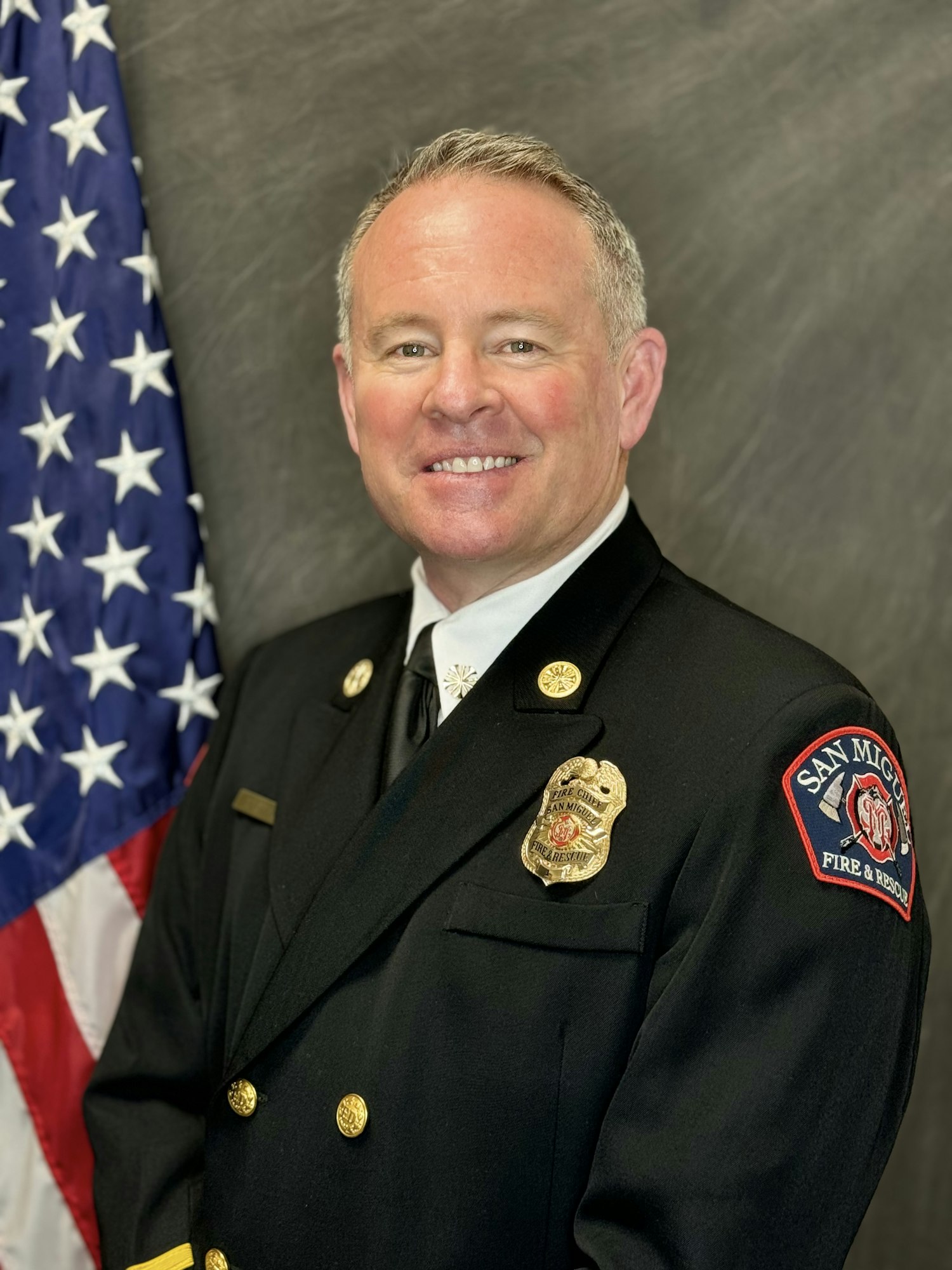 A smiling person in a formal firefighter uniform with badge, in front of an American flag.