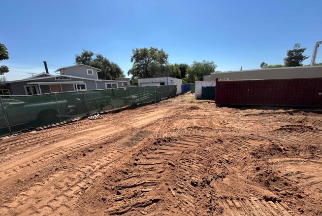 A construction site with bare soil, tire tracks, temporary fencing, portable toilets, and a dumpster under a clear sky.