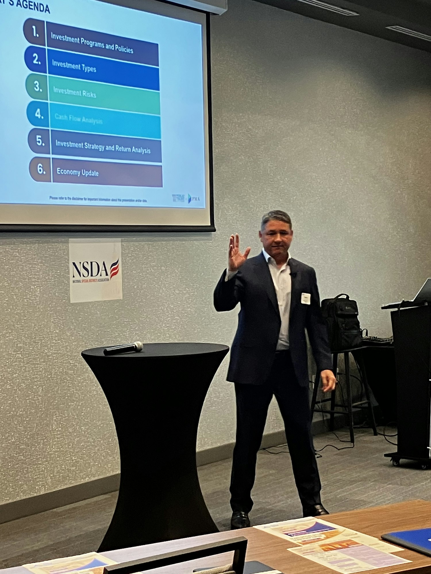A person in formal attire presenting with a slide titled "Today's Agenda" listing six topics related to investments.