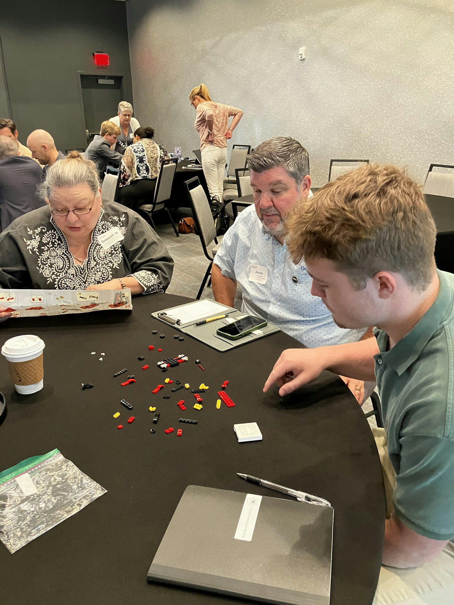 People sitting at a table assembling small Lego pieces with instructions, surrounded by papers and a coffee cup.