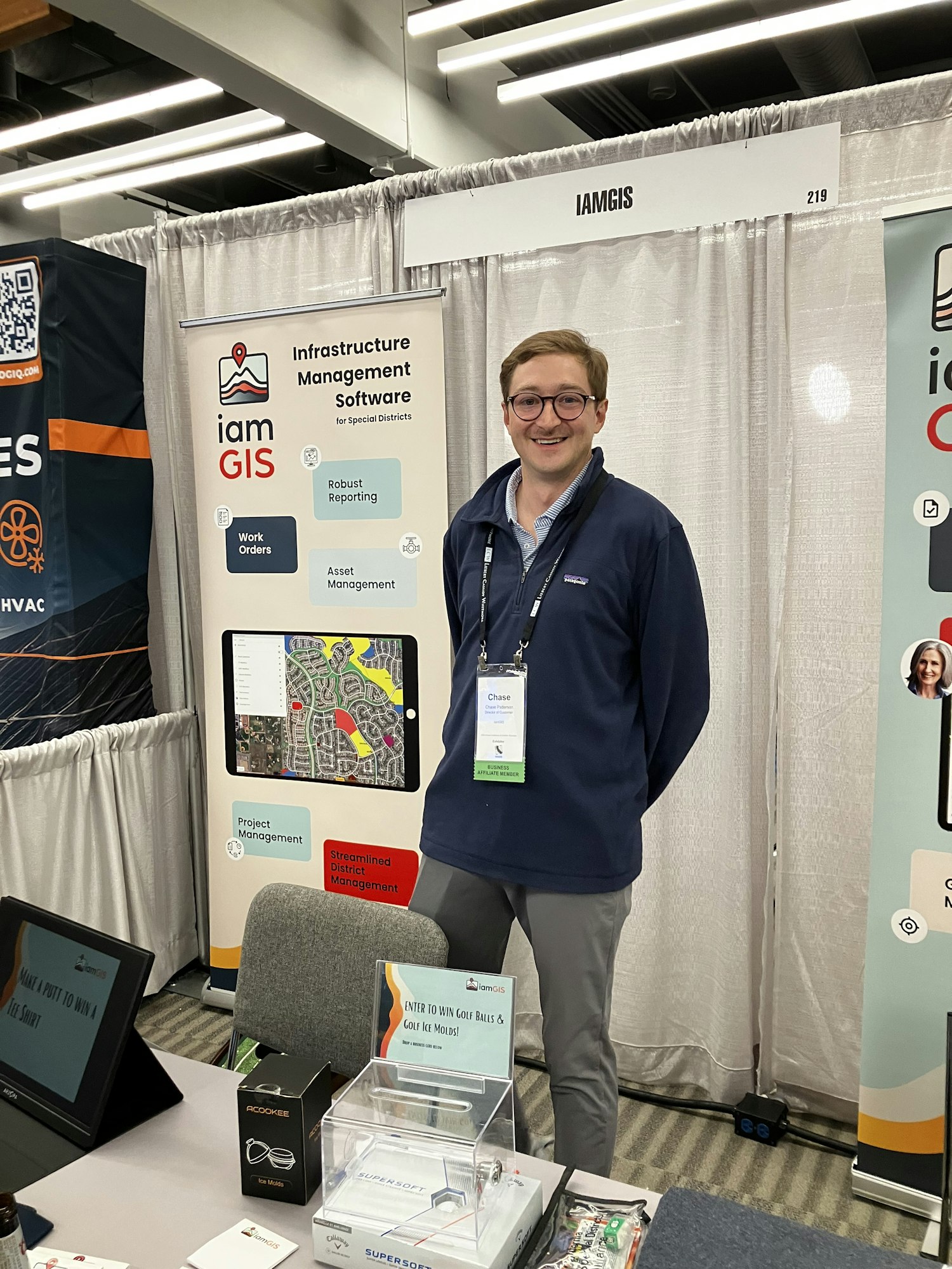 A person stands at an "IamGIS" booth featuring infrastructure management software at a convention or expo.