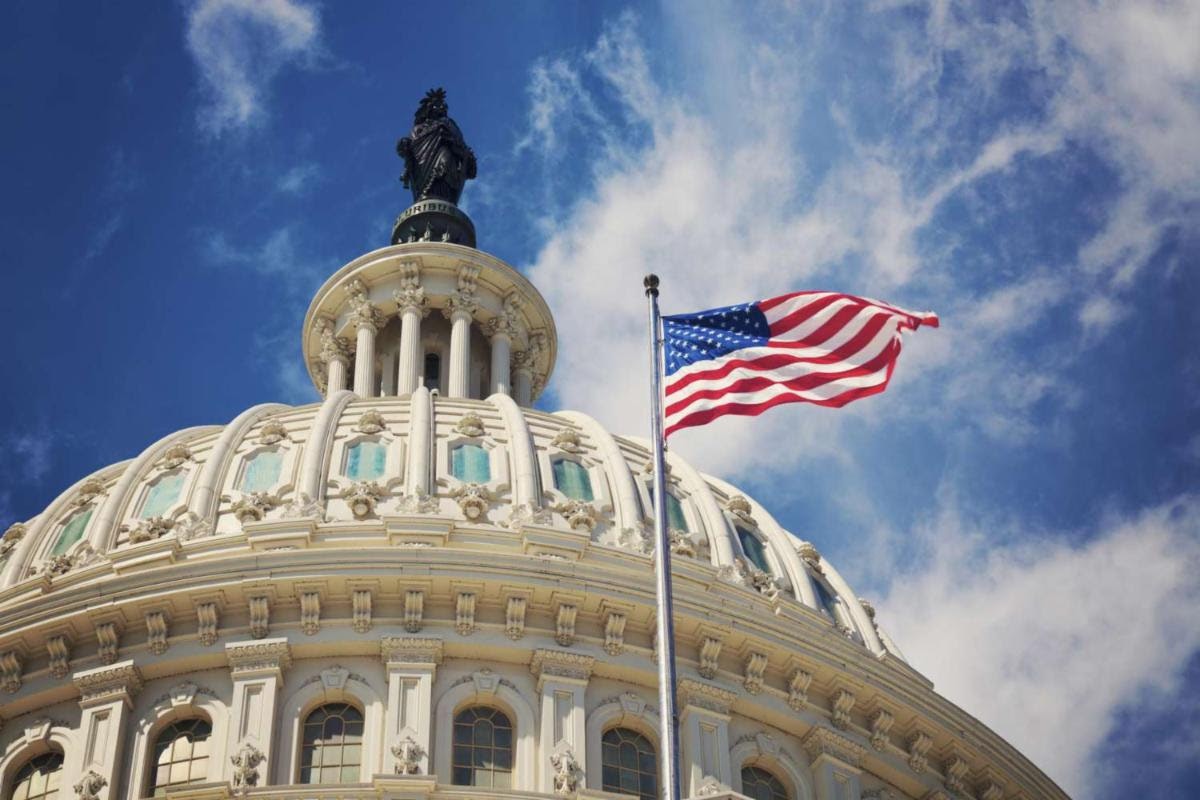 U.S. Capitol dome with an American flag waving in the foreground against a blue sky.
