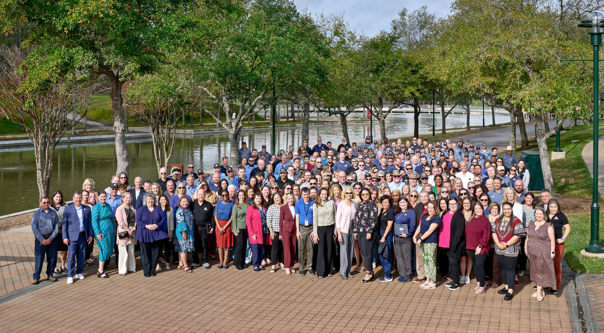 A large group of people posing for a photo outdoors, with trees and a waterway behind them.