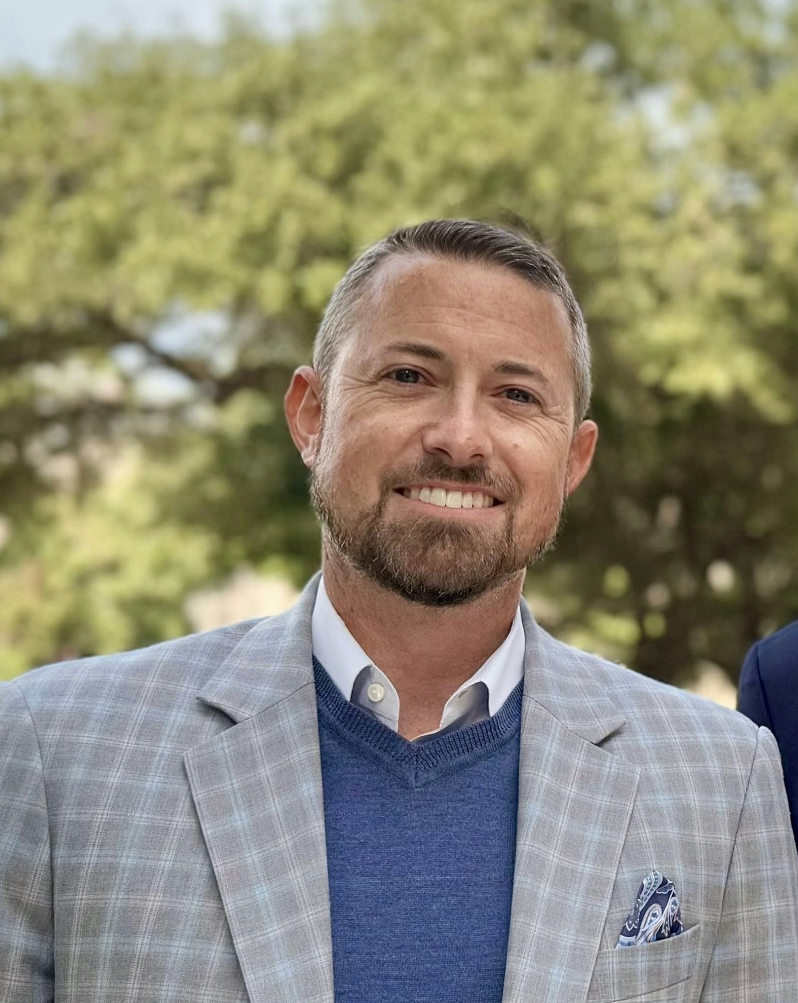 A man in a plaid jacket with a blue sweater, standing outdoors with a smile, against a blurry green background.