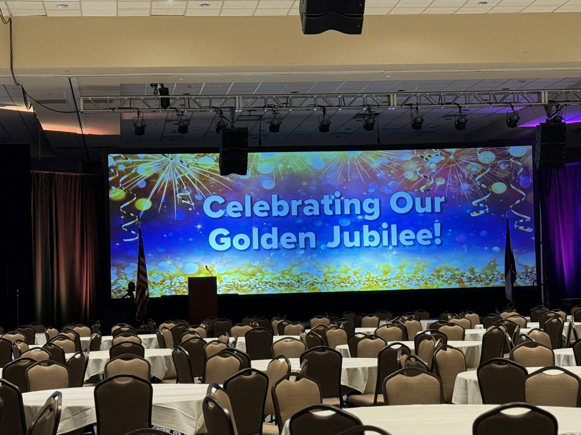 A conference room with decorated tables, chairs, and a stage displaying "Celebrating Our Golden Jubilee!"