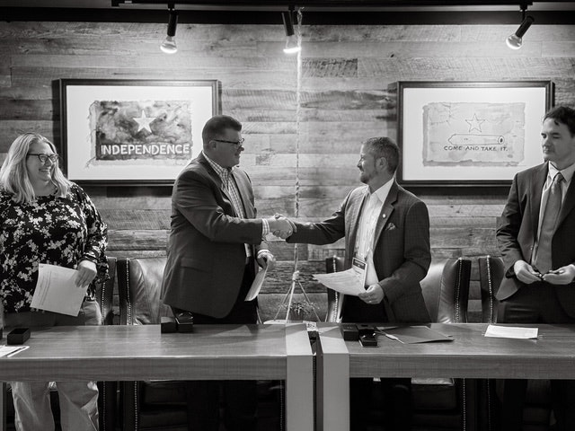 Four people at a table, two shaking hands, in a formal setting. Framed art on the wall.