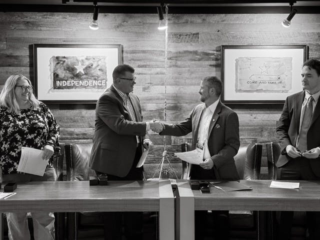 Four people at a table, two shaking hands, in a formal setting. Framed art on the wall.