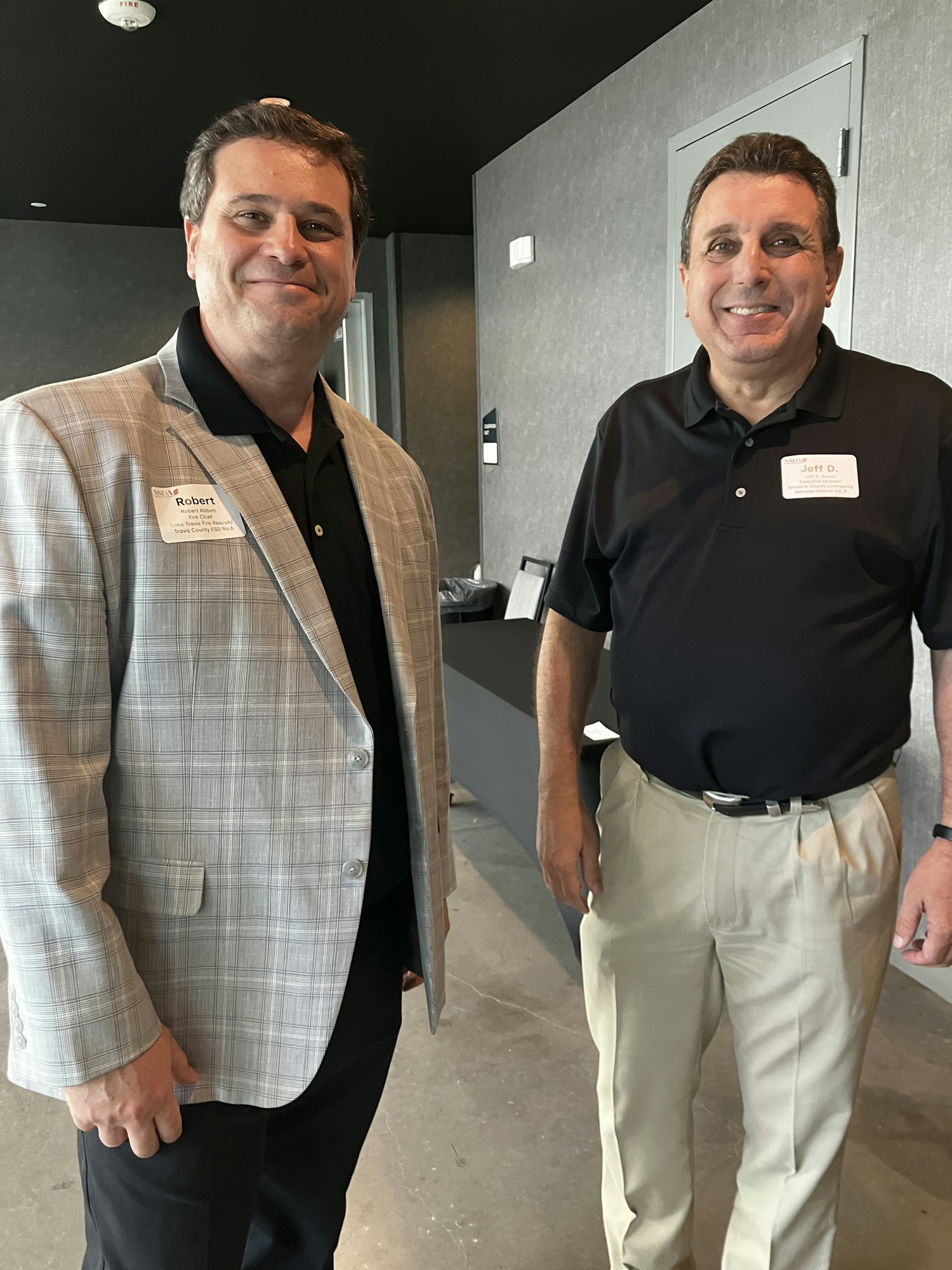 Two men smiling, wearing name tags, one in a black shirt and beige pants, the other in a checkered blazer.