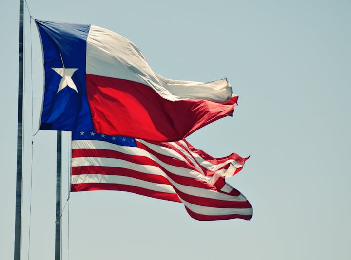 The Texas state flag and the United States flag are waving against a clear sky.