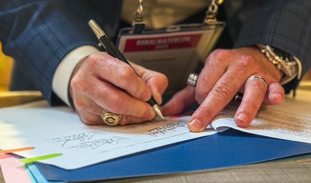 A person signing a document on a table, wearing rings and a lanyard.