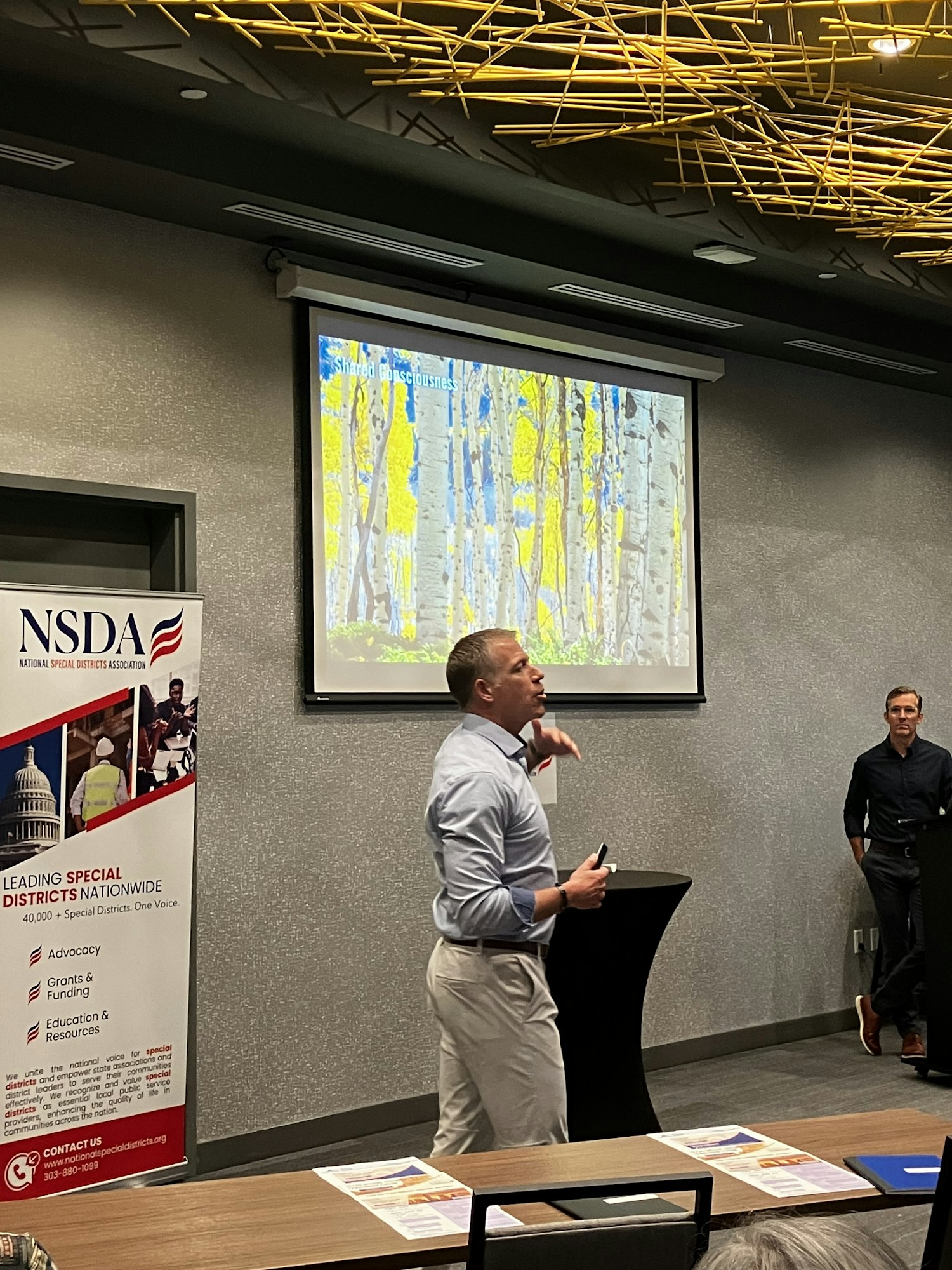 A person is speaking in front of a presentation screen with colorful visuals, and an NSDA banner is nearby.