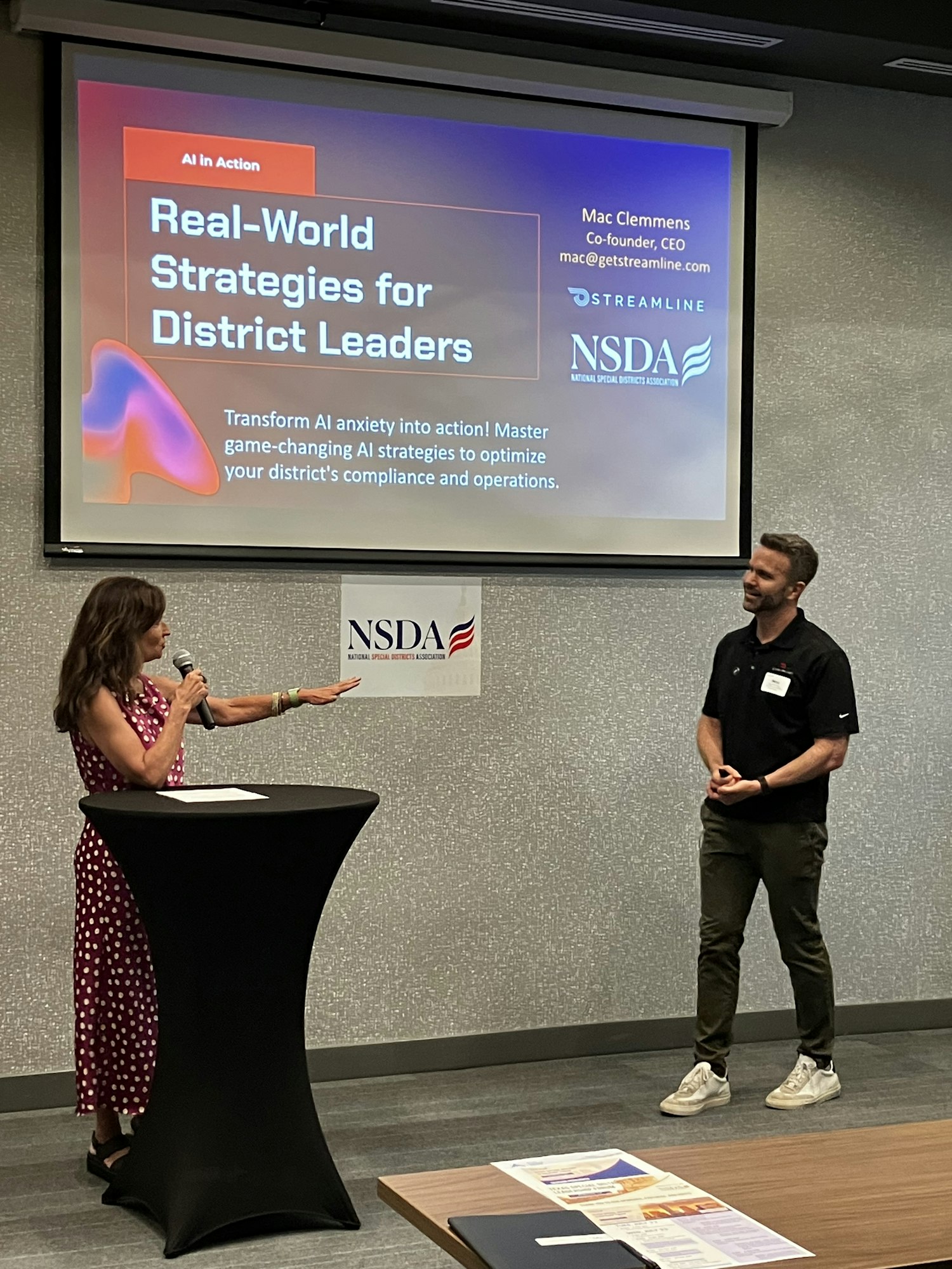 Presentation titled "Real-World Strategies for District Leaders" with a standing male speaker and seated female audience member.